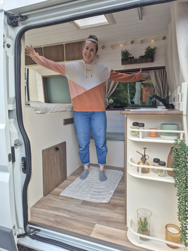 Femme souriante à l'entrée d'un van aménagé avec des meubles en bois sur mesure, idéal pour la vanlife et les voyages écologiques. Découvrez des aménagements de van fonctionnels et esthétiques avec Cosy Van à Strasbourg.